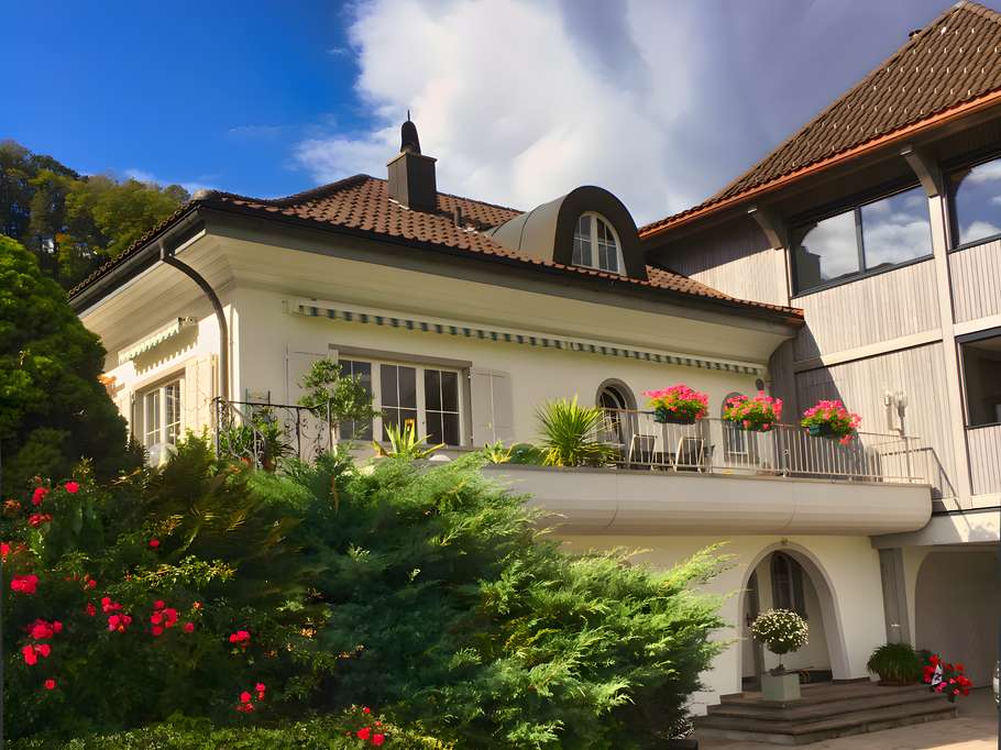 Zweistöckiges weißer Anbau mit dunklem Ziegeldach. Die große Terrasse oben ist mit rosafarbenen Blumenkästen, Gartenstühlen und einer Markise ausgestattet. Im Erdgeschoss führt eine Treppe zu einem bogenförmigen Eingang, umgeben von dichten grünen Hecken und blühenden Rosensträuchern.