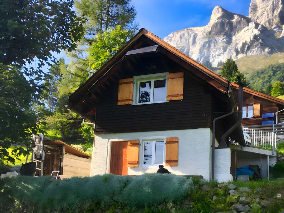 Kleines Berghaus mit dunkler Holzfassade und hellen Fensterläden vor hohen Felsen und Bäumen in einer sonnigen Berglandschaft.