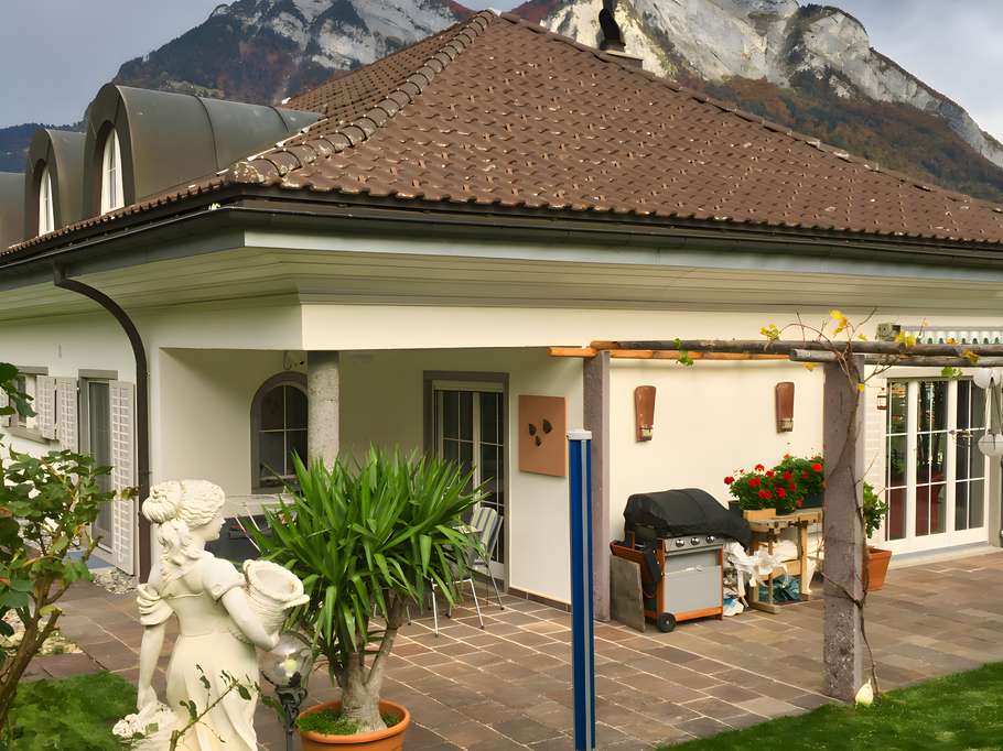 Außenansicht einer Terrasse mit Steinboden und angrenzender Rasenfläche. Zu sehen sind eine weiße Steinfigur im Vordergrund, eine Yucca-Palme im Topf, ein Gasgrill sowie eine hölzerne Pergola.