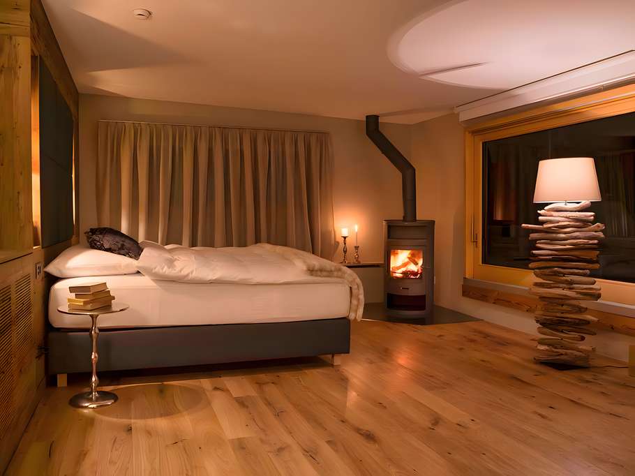 Helles, gemütlich eingerichtetes Schlafzimmer mit Doppelbett, Holzfußboden, Kaminofen und einer Stehlampe aus gestapelten Steinen neben dem Fenster.